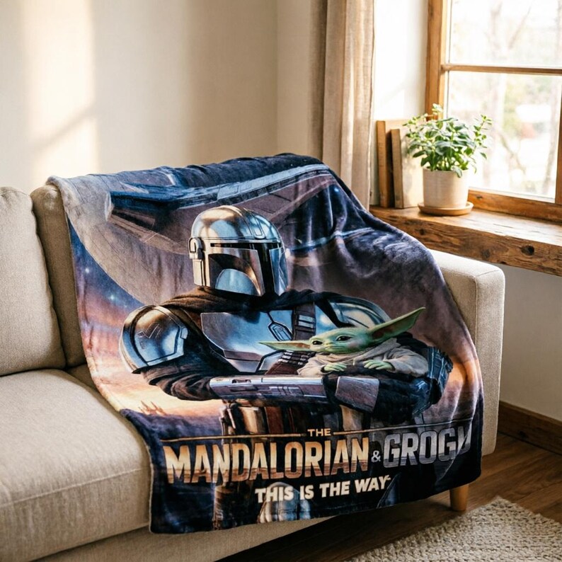 May include: A soft blanket featuring The Mandalorian and Grogu from the Star Wars series. The blanket displays the characters in a detailed graphic, with the text "THE MANDALORIAN & GROGU THIS IS THE WAY" at the bottom. The blanket is draped over a beige sofa.