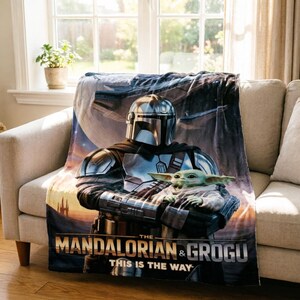 May include: A soft blanket featuring The Mandalorian and Grogu from the Star Wars series. The blanket shows the Mandalorian in his armour holding Grogu, with a space scene backdrop. The text "THE MANDALORIAN & GROGU" and "THIS IS THE WAY" are also visible.
