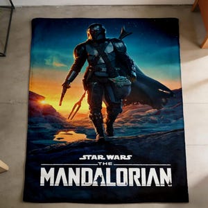 May include: A rectangular black blanket featuring a vibrant image of The Mandalorian from Star Wars. The artwork depicts the character in armour, holding a weapon, with a baby-like creature. The blanket has the text "STAR WARS THE MANDALORIAN".