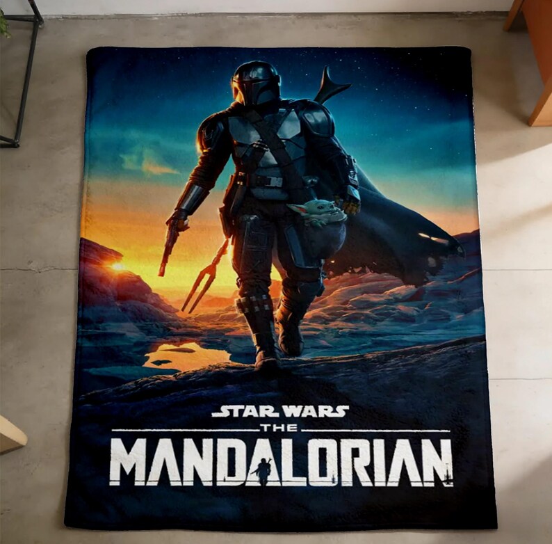 May include: A rectangular black blanket featuring a vibrant image of The Mandalorian from Star Wars. The artwork depicts the character in armour, holding a weapon, with a baby-like creature. The blanket has the text "STAR WARS THE MANDALORIAN".