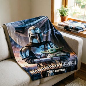 May include: A soft blanket featuring The Mandalorian and Grogu from the Star Wars series. The blanket displays the characters in a detailed graphic, with the text "THE MANDALORIAN & GROGU THIS IS THE WAY" at the bottom. The blanket is draped over a sofa.