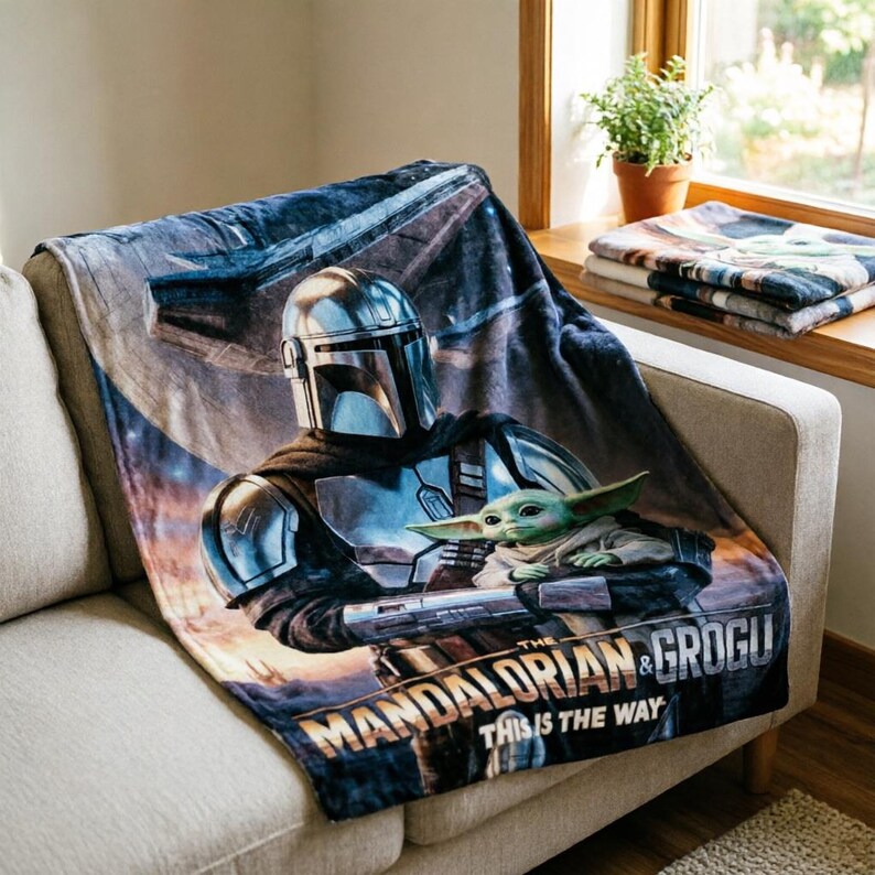 May include: A soft blanket featuring The Mandalorian and Grogu from the Star Wars series. The blanket displays the characters in a detailed graphic, with the text "THE MANDALORIAN & GROGU THIS IS THE WAY" at the bottom. The blanket is draped over a sofa.