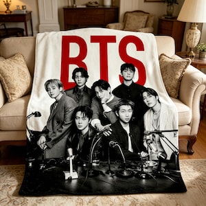 May include: A white blanket with the red text "BTS" and a group photo of seven men. The men are dressed in various outfits, and the blanket is draped over a beige sofa. The blanket has a black and white image of the group.
