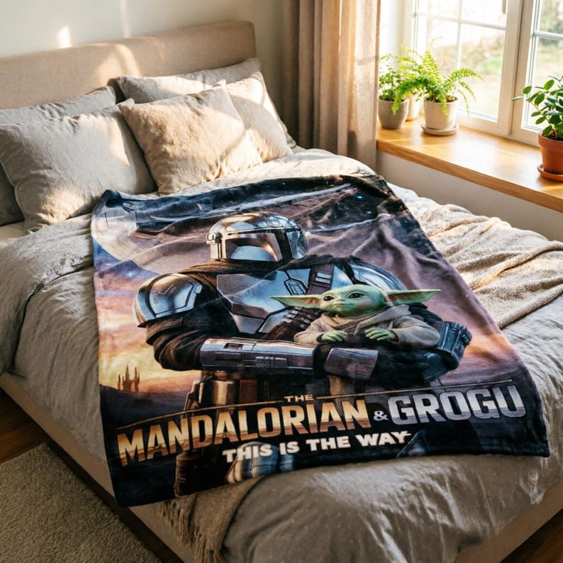 May include: A soft blanket featuring The Mandalorian and Grogu. The blanket shows a detailed image of the Mandalorian in silver armour holding Grogu. The text "The Mandalorian & Grogu" and "This is the Way" are printed on the blanket.