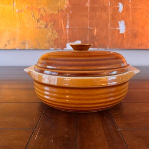 Puede incluir: Una cazuela de cerámica marrón con tapa. El plato tiene forma redonda con ranuras horizontales y asas pequeñas. La tapa tiene un pequeño pomo en la parte superior.