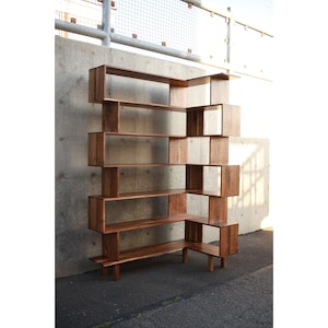 May include: A modern, geometric wooden bookshelf with multiple open shelves and cubby spaces. The bookshelf is constructed from dark-stained wood and features a unique, asymmetrical design. It stands against a concrete wall.