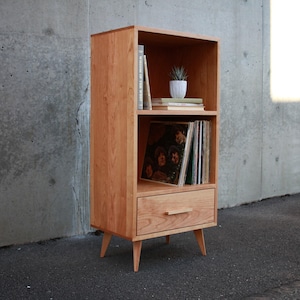 May include: A light brown wooden bookshelf with three shelves and a drawer. The top shelf holds books and a small potted plant. The middle shelf displays vinyl records. The bottom drawer has a wooden handle. The bookshelf is supported by four angled legs.