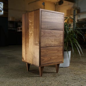 May include: A three-drawer wooden cabinet with a natural wood grain finish. The cabinet has a rectangular shape with angled legs. The drawers are flush with the cabinet's frame. A potted plant is next to the cabinet.