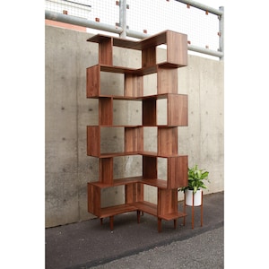 May include: A mid-century modern style wooden bookshelf with a unique, geometric design. The bookshelf is made of a warm brown wood and features multiple open shelves and cubbies. A small plant in a white pot sits nearby.