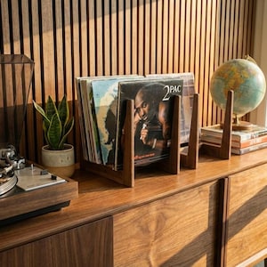 May include: A wooden record player with a vinyl record on the turntable. A wooden record holder displays several vinyl records, including one with a 2PAC album cover. A globe and books sit on a wooden cabinet.