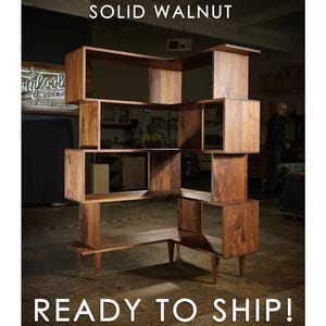 May include: A solid walnut, multi-level bookshelf with an open, geometric design. The unit features multiple cubby spaces and a unique, angular shape. Text at the top reads "SOLID WALNUT" and at the bottom "READY TO SHIP!"
