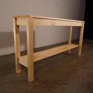 May include: A light-colored wooden console table with a rectangular top and a lower shelf. The table has four sturdy legs and is constructed from light-colored wood. The design is simple and modern, suitable for various home decor styles.