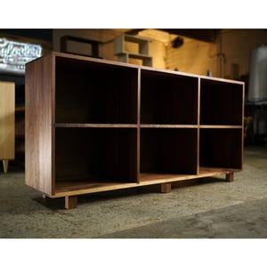 May include: A rectangular wooden bookshelf with six cubby holes. The bookshelf is made of dark brown wood and sits on short, light-colored wooden legs. The shelves are divided into two rows of three compartments each.