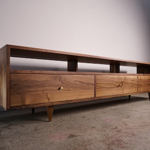 May include: A mid-century modern style media console made of wood. The console has two drawers and an open shelf. The drawers have brass knobs and the console has tapered legs. The wood has a warm, natural finish.