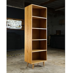 May include: A tall, light oak bookcase with five shelves and angled legs. The bookcase is made of solid wood and has a natural finish. The Tomlocker Wood Co. logo is visible in the background.