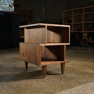 May include: A mid-century modern wooden side table with a two-tiered design. The table is made from dark brown wood and has a rectangular top and bottom with open storage. It stands on four angled legs. The dimensions are approximately 60 cm high.