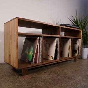 May include: Wooden record storage unit with multiple compartments, holding vinyl records. The unit is made of light and dark wood, with a rectangular shape and a low profile. The design is simple and modern, suitable for a living room or entertainment area.