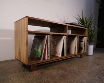 May include: Wooden record storage unit with multiple compartments, holding vinyl records. The unit is made of light and dark wood, with a rectangular shape and a low profile. The design is simple and modern, suitable for a living room or entertainment area.