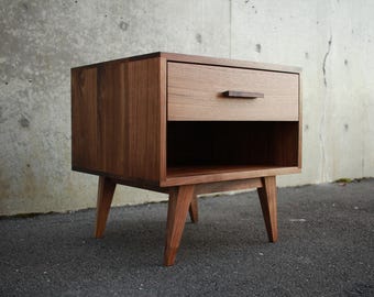 May include: A mid-century modern style wooden nightstand with a drawer and open shelf. The nightstand is made of a warm-toned wood and has tapered legs. The drawer has a simple, rectangular handle. The nightstand is approximately 24 inches tall.