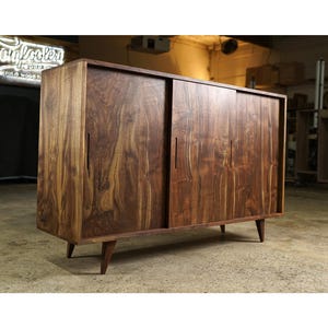 Peut inclure: Un buffet en bois avec deux portes coulissantes et quatre pieds inclinés. Le buffet est en bois brun foncé avec un motif de grain naturel. Le buffet a une forme rectangulaire et un design moderne du milieu du siècle.