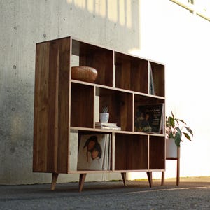 May include: A wooden bookcase with multiple compartments and shelves. The bookcase is made of light brown wood and has a mid-century modern design. It contains decorative items such as a wooden bowl, books, and vinyl records. A small potted plant sits on the side.