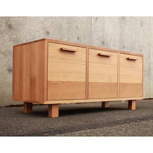May include: A light brown wooden cabinet with three doors and a rectangular shape. The cabinet has a natural wood grain finish and is elevated on short, square legs. Each door features a simple, horizontal handle.