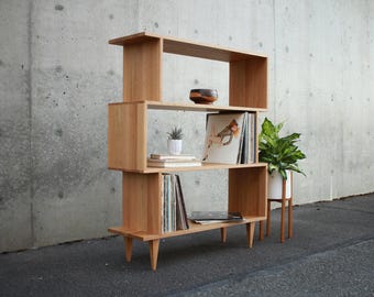 May include: A mid-century modern wooden bookshelf with a unique, staggered design. The light-colored wood shelf holds vinyl records, books, and decorative items. A potted plant and a small succulent add a touch of greenery to the display.