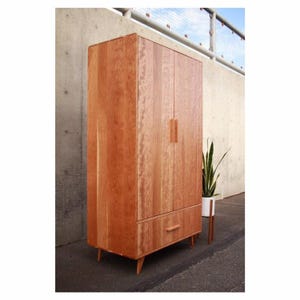 May include: A tall, mid-century modern wooden wardrobe with a warm, reddish-brown finish. The wardrobe has two doors, a drawer, and tapered legs. The doors have vertical wooden handles. A potted plant sits beside the wardrobe.