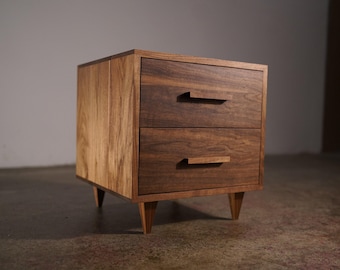 May include: A two-drawer wooden nightstand with a mid-century modern design. The nightstand is constructed from two types of wood, with a lighter wood on the sides and a darker wood on the drawers. The drawers have rectangular handles and are supported by angled legs.