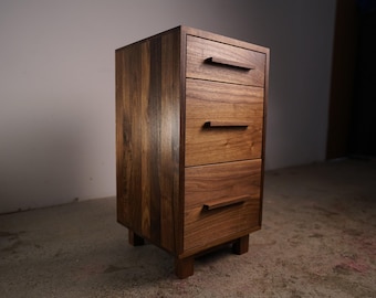 May include: A small, three-drawer wooden cabinet with a warm, brown finish. The cabinet has a rectangular shape with a simple design, featuring horizontal drawer pulls. It stands on short, square legs.