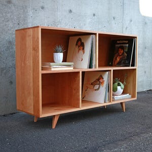 May include: A mid-century modern style wooden record shelf with six compartments. The shelf is made of light-colored wood and has angled legs. Records and small potted plants are displayed inside the shelf. The background is a concrete wall.