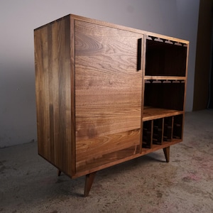 May include: A wooden drinks cabinet in a warm, natural finish. The cabinet has a door on the left, a section for wine glasses, and a lower section for wine bottles. It stands on angled legs, offering a mid-century modern aesthetic.