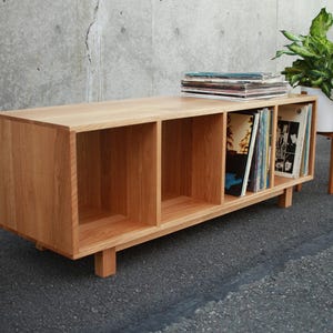 May include: A light-colored wooden record storage cabinet with four open compartments. The cabinet is rectangular and sits on short legs. Vinyl records are visible in two compartments. A stack of records sits on top.