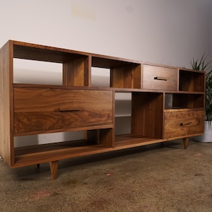 May include: A long, low-profile wooden media console crafted from dark walnut wood. The unit features a combination of open shelving compartments and three drawers with horizontal wooden handles, all supported by four short, tapered wooden legs.