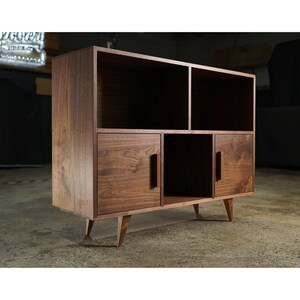 May include: A mid-century modern style wooden cabinet with a warm brown finish. The cabinet features open shelving, two doors, and a central open compartment. It stands on angled wooden legs, offering a stylish storage solution.