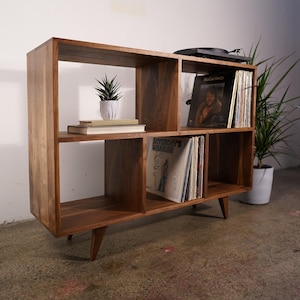 May include: A mid-century modern wooden record shelf with multiple compartments. The shelf holds vinyl records, books, and a small potted plant. The shelf is made of a warm-toned wood and has angled legs.