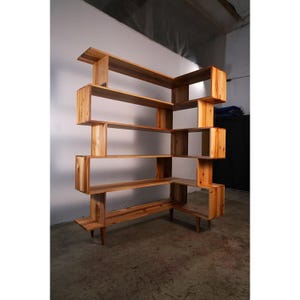 May include: A wooden bookshelf with a unique, geometric design. The shelves are arranged in an asymmetrical pattern, with some sections extending outward. The natural wood finish gives it a warm, inviting look. The bookshelf is supported by thin legs.