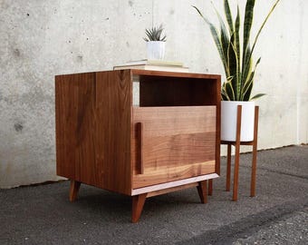 May include: A mid-century modern style wooden nightstand with a single door and open shelf. The nightstand is made of dark brown wood and has tapered legs. A small white potted plant and books sit on top. A white plant pot with a tall green plant is next to it.