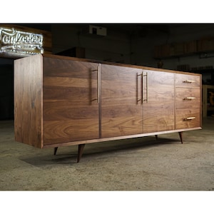 May include: A mid-century modern style wooden cabinet with a rich brown finish. The cabinet features two doors, three drawers, and brass-colored handles. The cabinet is supported by angled wooden legs. The sign in the background reads "Tomfoolery Woods".