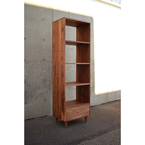 May include: A tall, wooden bookshelf in a warm, reddish-brown finish. It has four open shelves and a bottom drawer with a small, round wooden knob. The bookshelf stands on four tapered legs.