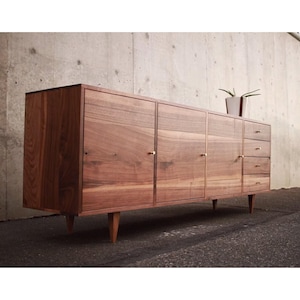 May include: A mid-century modern wooden cabinet with four doors and three drawers. The cabinet is made of a warm-toned wood and features brass hardware. The cabinet is supported by four tapered legs. A small potted plant sits on top.