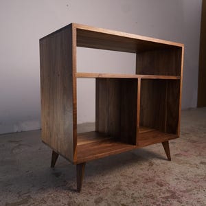 May include: A wooden bookshelf in a mid-century modern style with a natural finish. It features a rectangular design with open compartments and tapered legs. The visible wood grain enhances its aesthetic appeal.