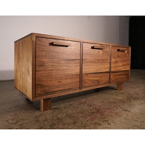 Peut inclure: Un meuble en bois avec trois tiroirs et une porte, tous avec une finition en grain de bois naturel. Le meuble repose sur de courts pieds carrés. Les poignées sont encastrées dans le bois, créant un look élégant et moderne. Le bois a une teinte marron chaude.