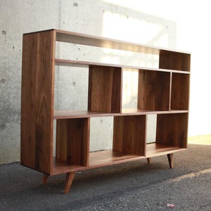 May include: A mid-century modern style wooden bookshelf with multiple compartments and shelves. The bookshelf is made of dark brown wood and has a rectangular shape with angled legs. The design features open storage spaces of varying sizes.