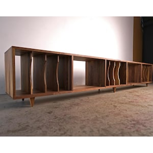 May include: A long, rectangular wooden media console with multiple open compartments. The console is made of a warm-toned wood and features a series of vertical dividers and angled legs. The design is mid-century modern, suitable for storing records or media.