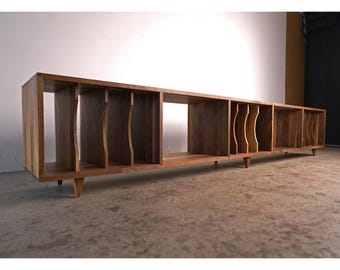 May include: A long, rectangular wooden media console with multiple open compartments. The console is made of a warm-toned wood and features a series of vertical dividers and angled legs. The design is mid-century modern, suitable for storing records or media.