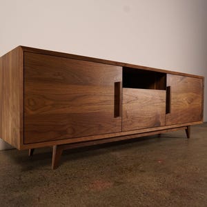 May include: A mid-century modern style wooden media console. The cabinet is made of a warm-toned wood with visible grain. It features two doors, a central open compartment, and a drawer. The console is supported by angled legs.