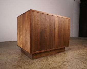 May include: A wooden cabinet with two doors and a rectangular base. The cabinet is made of dark brown wood with visible grain and a natural finish. The doors are flush with the frame, and the cabinet sits on a low, rectangular base.