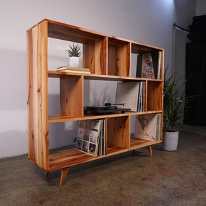 May include: A wooden bookshelf with multiple cubbyhole shelves. The natural wood-toned bookcase has a mid-century modern design with angled legs. A record player, vinyl records, and a small potted plant are visible on the shelves.