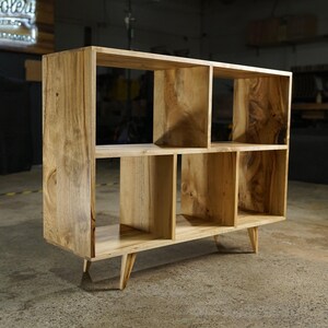 May include: A light-colored wooden bookshelf with six cubby spaces. The bookshelf is rectangular with a natural wood grain finish and sits on four angled legs. The design is modern and minimalist, suitable for storage or display.